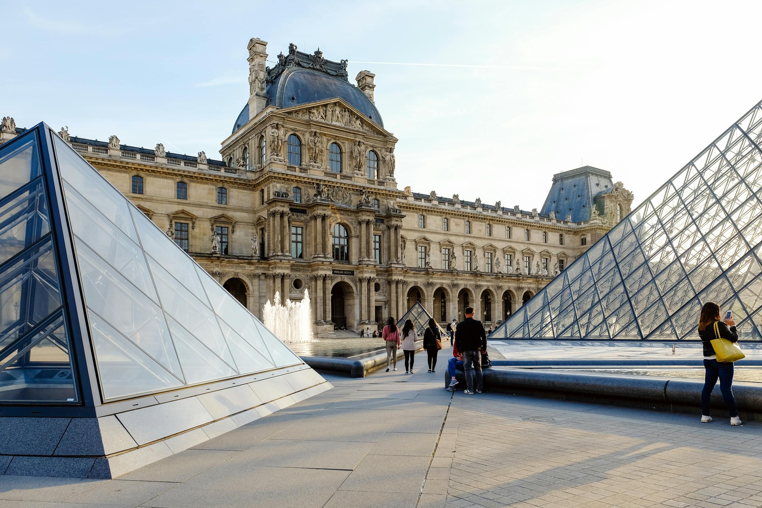 The Louvre Museum and its glass pyramids in Paris, bustling with tourists on a sunny day.